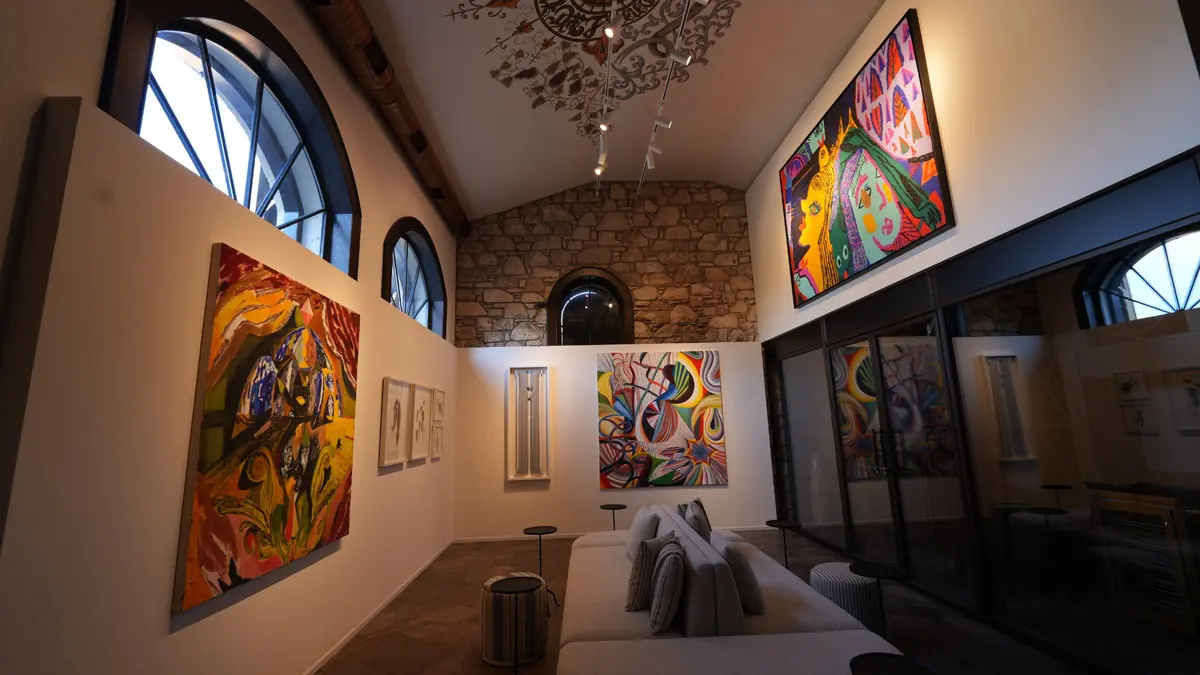 Yüksek tavanlı, kemerli pencereleri ve rahat bir oturma alanı bulunan modern bir sanat galerisinin genel görünümünde, duvarlarda sergilenen çeşitli renkli ve soyut eserler, LS Contemporary Alaçatı'daki karma serginin canlı atmosferini yansıtıyor.