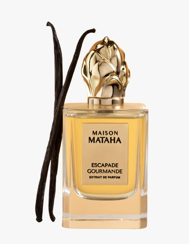 Maison Mataha Escapade Gourmande Extrait de Parfum şişesi, beyaz bir arka plan önünde, altın rengi, yaprak desenli kapak ve etiketiyle sergileniyor, şişenin yanında duran iki koyu renkli vanilya çubuğu ise kokunun gurme ve tatlı profilini vurguluyor.