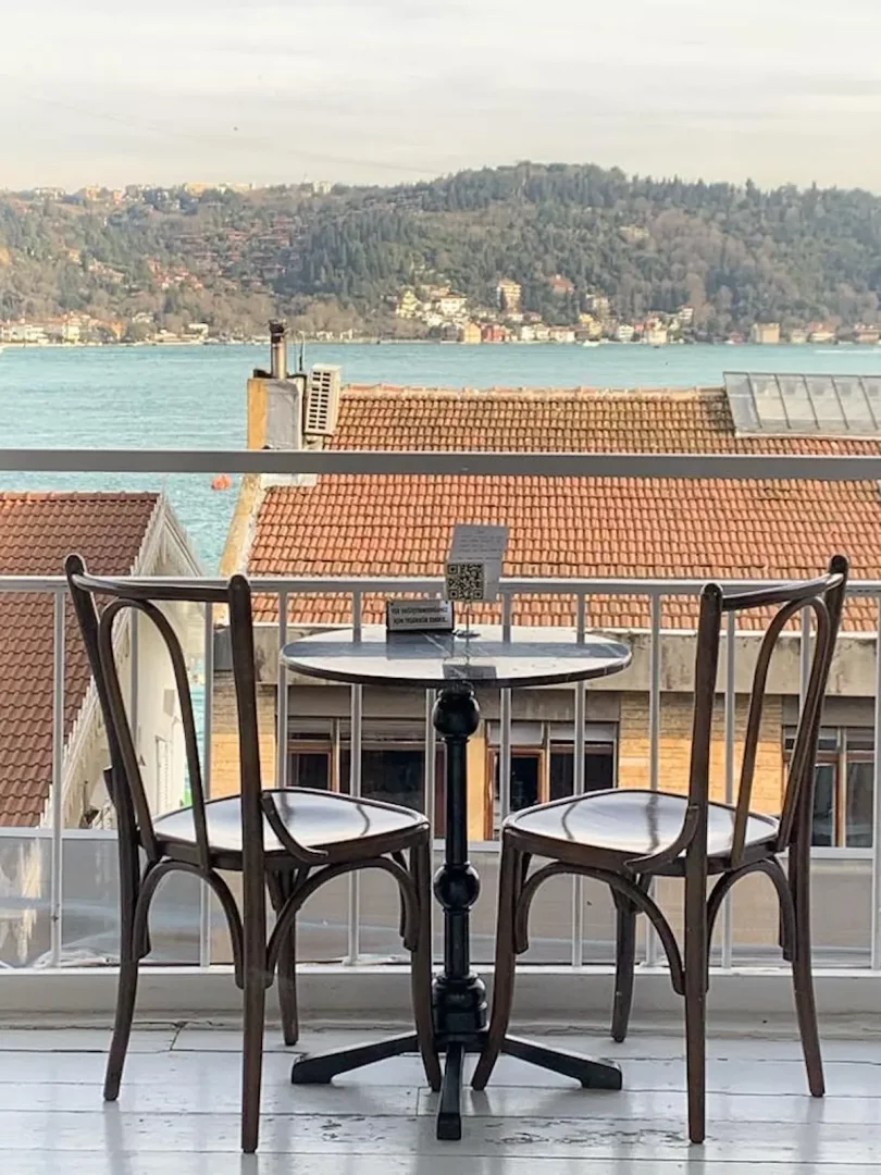 Bebek'teki ünlü bir kafe balkonunda, iki ahşap sandalye ve üzerinde QR kodlu menü bulunan yuvarlak bir masanın önünden görünen, Boğaz'ın masmavi suları, karşı kıyıdaki yemyeşil tepeler ve yerleşim alanlarının oluşturduğu büyüleyici İstanbul manzarası.