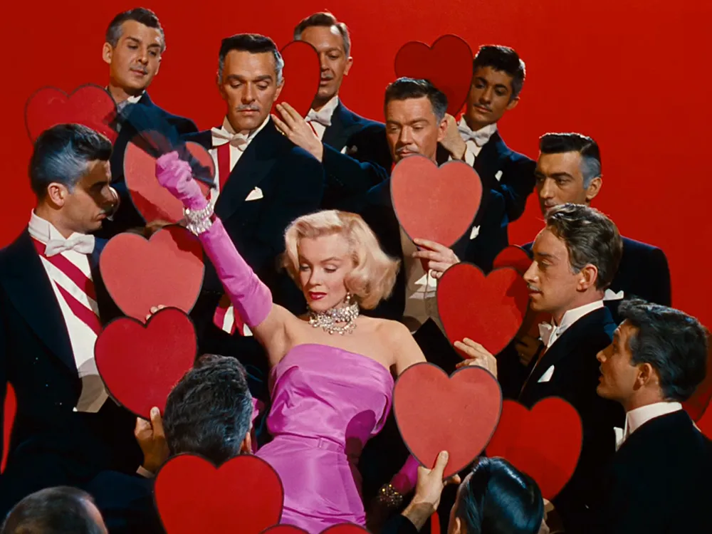 Pembe straplez elbisesi, uzun pembe eldivenleri ve pırlanta kolyesiyle göz kamaştıran Marilyn Monroe, onu hayranlıkla çevreleyen smokinli beylerin uzattığı kırmızı kalp figürleri arasında, Erkekler Sarışın Sever filminin ikonik bir sahnesinde adeta ışık saçıyor.