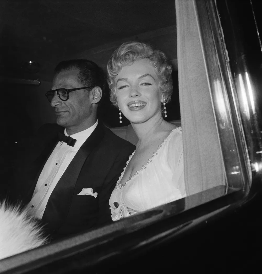Marilyn Monroe ve Arthur Miller, bir arabanın arka koltuğunda, Monroe'nun beyaz elbisesi ve inci küpeleriyle kameraya gülümserken, Miller'ın ise gözlüklü ve smokinli bir şekilde yana baktığı, ünlü evliliklerinden samimi bir siyah beyaz an.