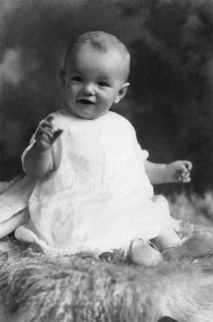 Siyah beyaz bir fotoğrafta, çocukluğundan bir karede, kameraya doğru içtenlikle gülümseyen ve beyaz, kabarık bir elbise giymiş bebek Marilyn Monroe, tüylü bir kilim üzerinde otururken sağ elini sevimli bir şekilde kaldırmış, gelecekteki yıldızın ilk yıllarına dair sıcak bir anı yansıtıyor.