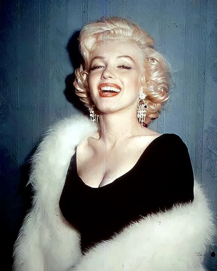 Marilyn Monroe kimdir içeriğine gönderme yapan bu görselde, açık sarı kıvırcık saçları, neşeli ve ışıltılı gülümsemesi, büyük parlak küpeleri, derin dekolteli siyah elbisesi ve omuzlarındaki kabarık beyaz kürküyle kameraya bakarken çekilmiş ikonik bir fotoğrafı görülüyor.