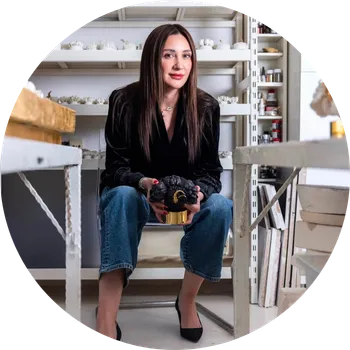 Sanatçı Melis Buyruk, kendi sanat stüdyosunda koyu renk kadife ceketi, kot pantolonu ve topuklu ayakkabılarıyla oturmuş, ellerinde altın rengi bir kaide üzerinde duran koyu renk, organik formda bir heykeli sergilerken, arkasında raflarda sayısız küçük beyaz heykeller ve sanat malzemeleri görülüyor.