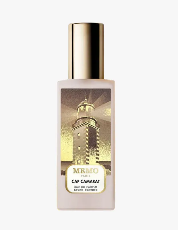 Ortasında Memo Paris markasına ait Cap Camarat Eau de Parfum yazan, üzerinde ışık saçan bir deniz feneri illüstrasyonunun altın rengi tonlarında yer aldığı, dikdörtgen buzlu cam şişede ve altın renkli kapaklı bir parfüm şişesi.