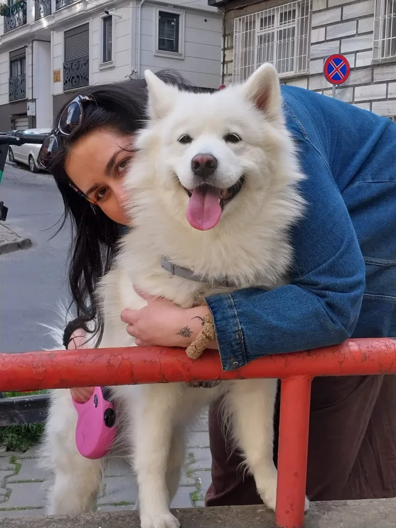 Uzun koyu saçlı, kot ceketli bir kadın, yüzünün bir kısmı beyaz kabarık tüylü, dili dışarıda gülümseyen ve kameraya bakan köpeğinin arkasından kırmızı bir metal korkuluğa yaslanmış, köpeğine sarılırken, pembe tasması ve arka planda şehir binaları ile park yapılmaz işareti net bir şekilde görülüyor.