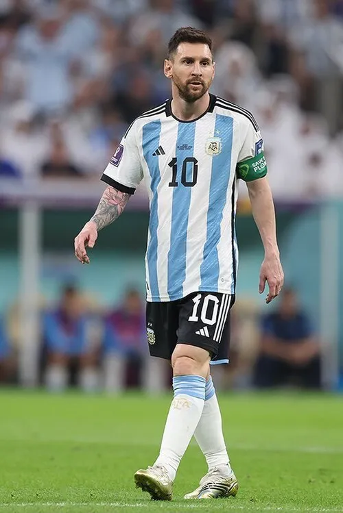 2022 Dünya Kupası'nda Arjantin Milli Takımı'nın beyaz ve açık mavi çizgili 10 numaralı formasını, siyah şortunu ve beyaz çoraplarını giyen Lionel Messi, yeşil bir futbol sahasında, seyircilerin olduğu bulanık bir stadyum arka planında sakin bir şekilde duruyor. Sol kolunda ise Save Plant yazılı yeşil bir pazu bandı ve ayağında altın rengi kramponlar var.