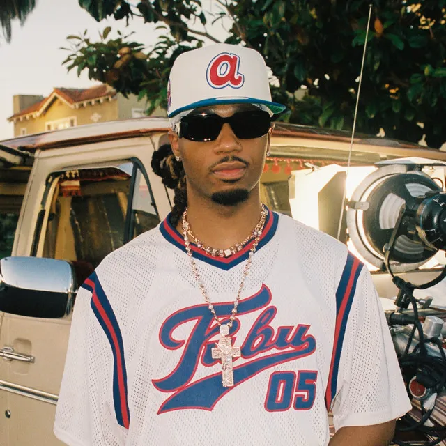 Metro Boomin, beyaz şapka, güneş gözlüğü ve Fubu yazılı beyaz forma giyerek, boynunda haç kolye ve zincirleriyle, dış mekanda bir çekimde modern hip-hop ve trap sahnesinin en etkili prodüktörlerinden biri olarak karizmatik bir duruş sergiliyor.