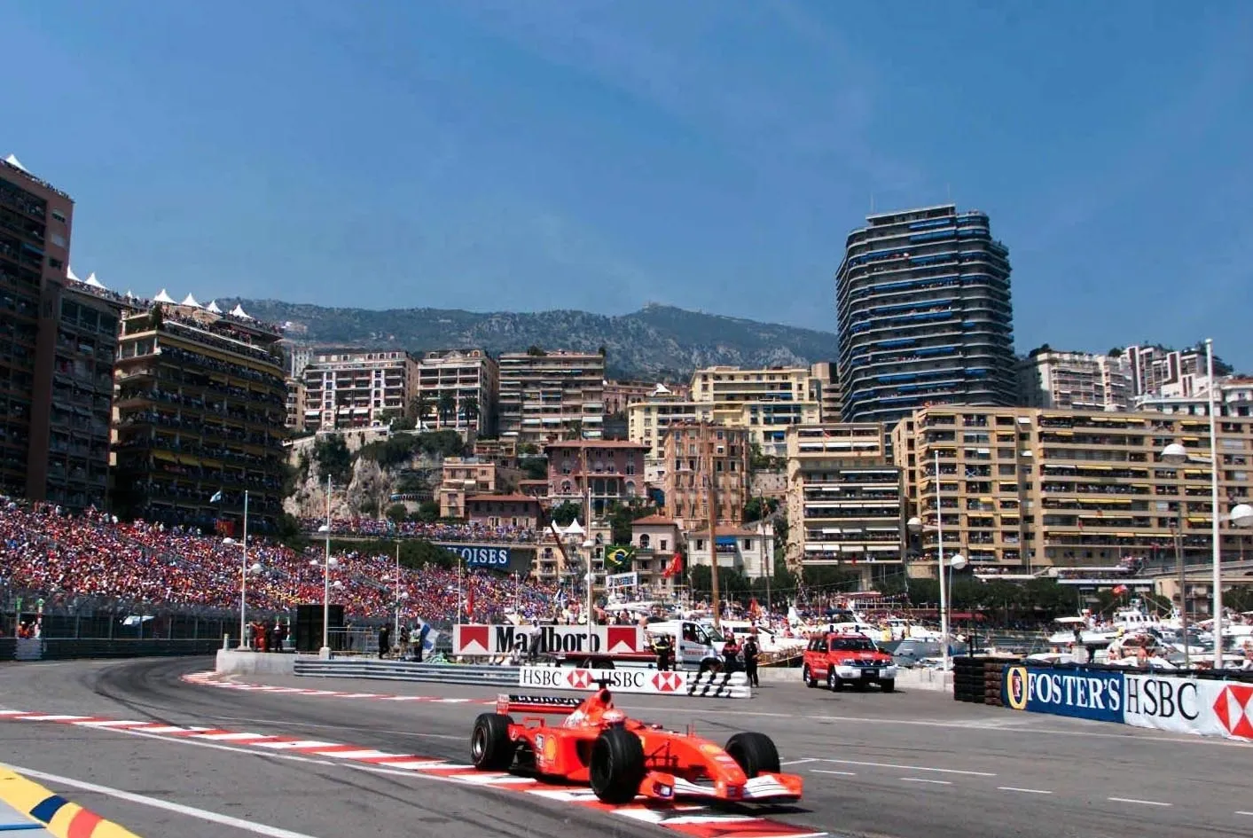 2001 Monako Grand Prix'sinde, Michael Schumacher'in sürdüğü kırmızı Ferrari Formula 1 aracı, şehrin lüks binaları, kalabalık tribünler ve dağlık bir arka plan eşliğinde, Marlboro ve HSBC reklam panolarının çevrelediği keskin bir virajdan geçerken gösteriliyor.