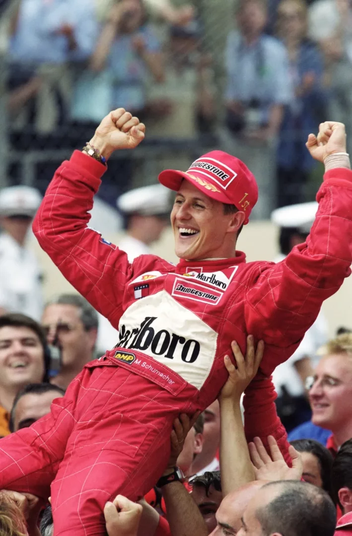 Michael Schumacher, 2001 Monaco GP zaferini kutlarken, kırmızı yarış tulumu ve şapkasıyla, gülümseyerek havaya kaldırdığı yumruklarıyla neşeli bir kalabalık tarafından omuzlarda taşınıyor; etrafındaki coşkulu yüzler onun başarısını paylaşıyor.