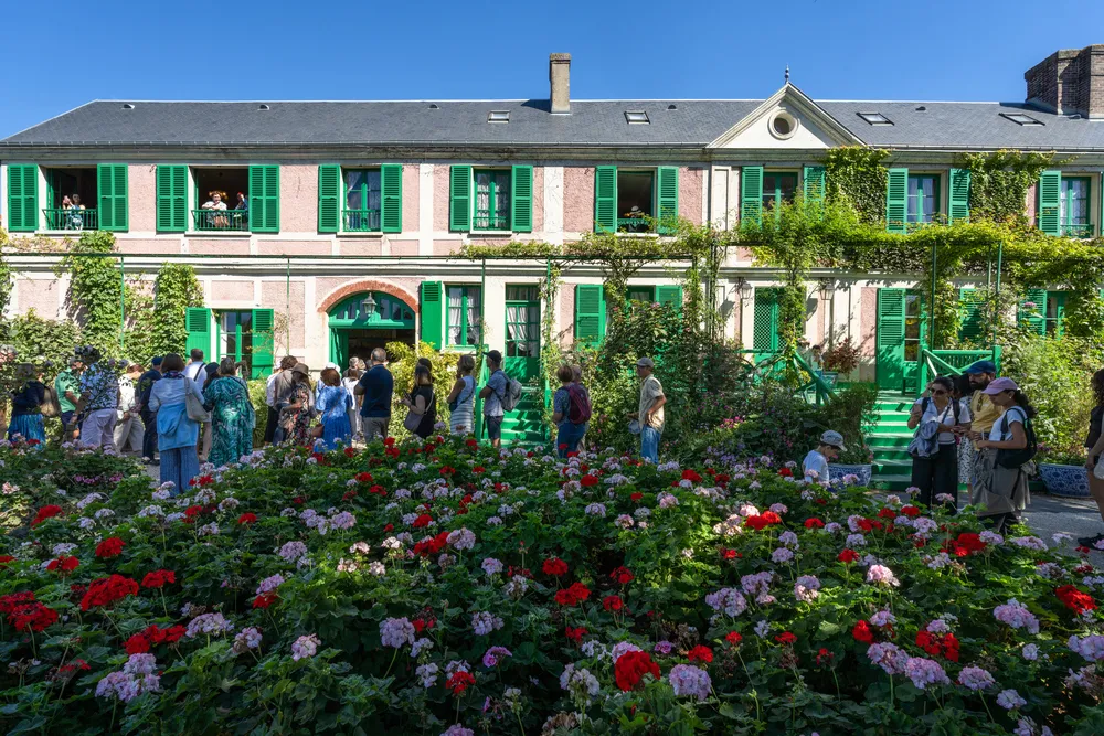 Güneşli bir günde, Fransız ressam Claude Monet'nin Giverny'deki pembe cepheli, yemyeşil panjurlu evi, ön kısmında açmış kırmızı ve pembe sardunyalarla dolu bakımlı çiçek bahçesini ziyaret eden kalabalık insanlarla birlikte göz alıcı bir şekilde betimlenmiştir.