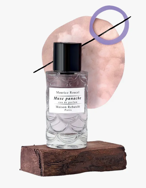 Koyu renkli, pürüzlü ahşap bir bloğun üzerinde duran, alt kısmında dalgalı desenlere sahip, şeffaf cam şişedeki Maurice Roucel imzalı Musc panache eau de parfum, arkasında pembe-kahverengi soyut bir şekil, ince siyah bir çizgi ve mor bir halka ile sanatsal bir kompozisyon oluşturuyor.