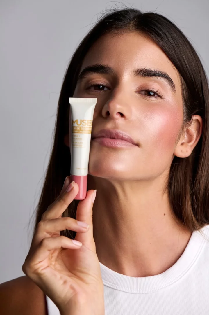 Yanaklarına uyguladığı doğal kiraz krem allık ile sağlıklı bir ışıltıya sahip, uzun koyu saçlı bir kadın, beyaz bir tişört giymiş ve elinde üzerinde Muse Lab markalı Cheek to Cheek Cherry Cream Blush yazan küçük bir allık tüpü tutuyor.