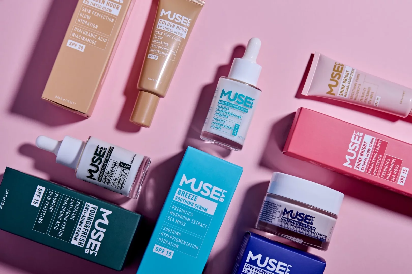 Pembe bir zemin üzerinde dağınıkça yerleştirilmiş Muse Lab cilt bakım ürünleri koleksiyonu, farklı renklerdeki kutu ve şişeleriyle dikkat çekiyor; SPF korumalı Golden Hour BB Tinted Glow, Breeze Soothing Serum ve Young & Fresh Anti-Aging Serum gibi çeşitli ürünler arasında, hyaluronik asit ve prebiyotik gibi aktif içerikler vurgulanıyor.