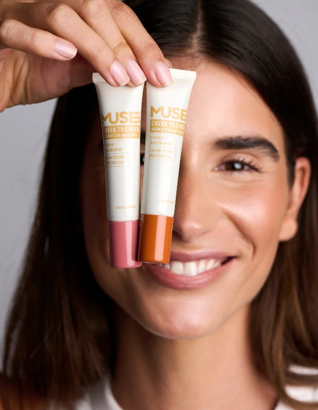 Mutlu ve esmer tenli bir kadın, gözlerinin önünde Muse Labs Cheek to Cheek markasına ait pembe tonlu Berry Cream Blush ve bronz tonlu Bronze Cream Blush ürünlerini tutarak kocaman gülümserken, yanaklarında bu nemlendirici krem allıkların verdiği sağlıklı ve doğal ışıltıyı sergiliyor.