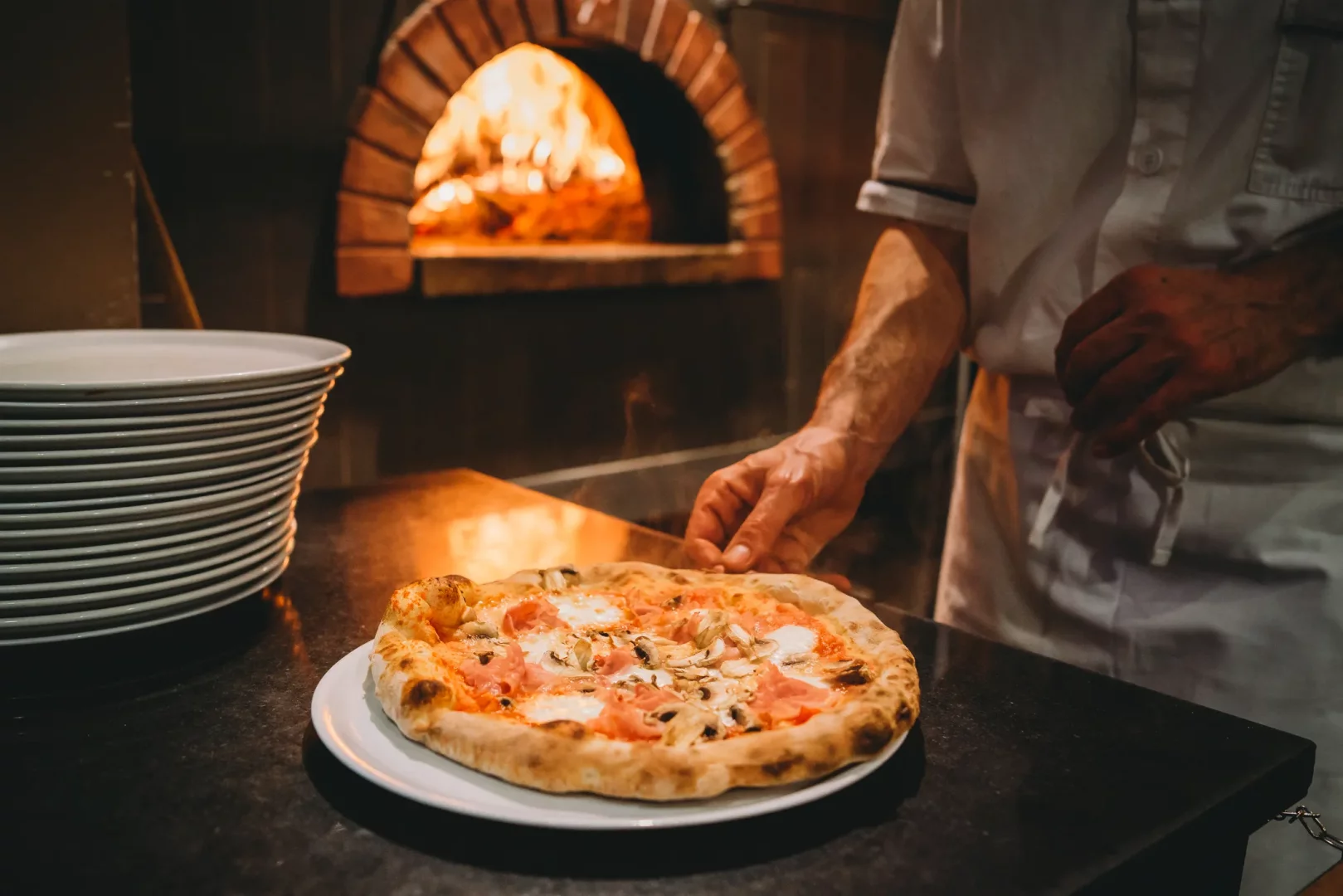 Bir şef, odun ateşinde taş fırından yeni çıkmış, jambon, mantar ve mozzarella peyniri ile hazırlanan, beyaz bir tabağın üzerindeki buharda tüten bir Napoliten pizzayı tezgaha yerleştirirken, solda temiz tabak yığını ve arka planda alevli fırın görünüyor.
