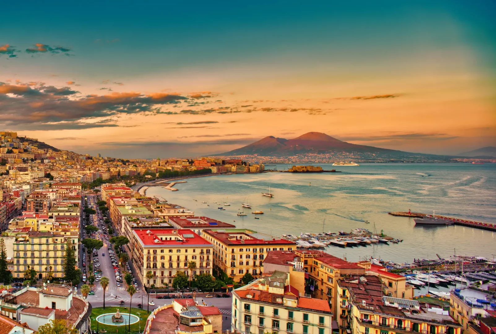 Napoli'nin binalarla dolu kıyı şeridinin, hareketli ana caddelerin, teknelerle dolu masmavi körfezin ve Castel dell'Ovo gibi tarihi yapıların, ufukta yükselen etkileyici Vezüv Yanardağı ile gün batımının sıcak tonlarıyla aydınlandığı panoramik bir görüntüsü.