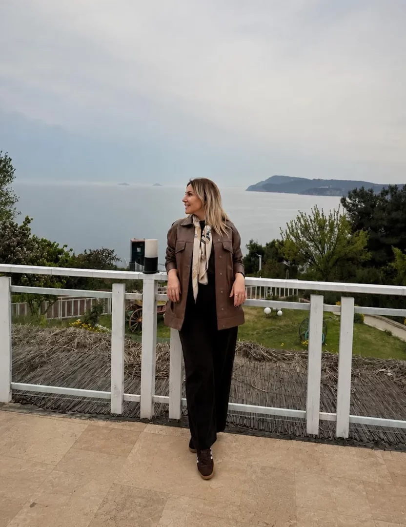 Nazan Zorlu, kahverengi bir ceket, açık renk desenli bir şal ve koyu renk bol paça pantolon giymiş halde, deniz manzaralı geniş bir terasta beyaz ahşap korkuluklara yaslanmış, bulutlu gökyüzünün altında uzaklara ve denize doğru huzurla gülümseyerek bakıyor, arka planda yeşil ağaçlar ve uzaktaki adalar seçiliyor.