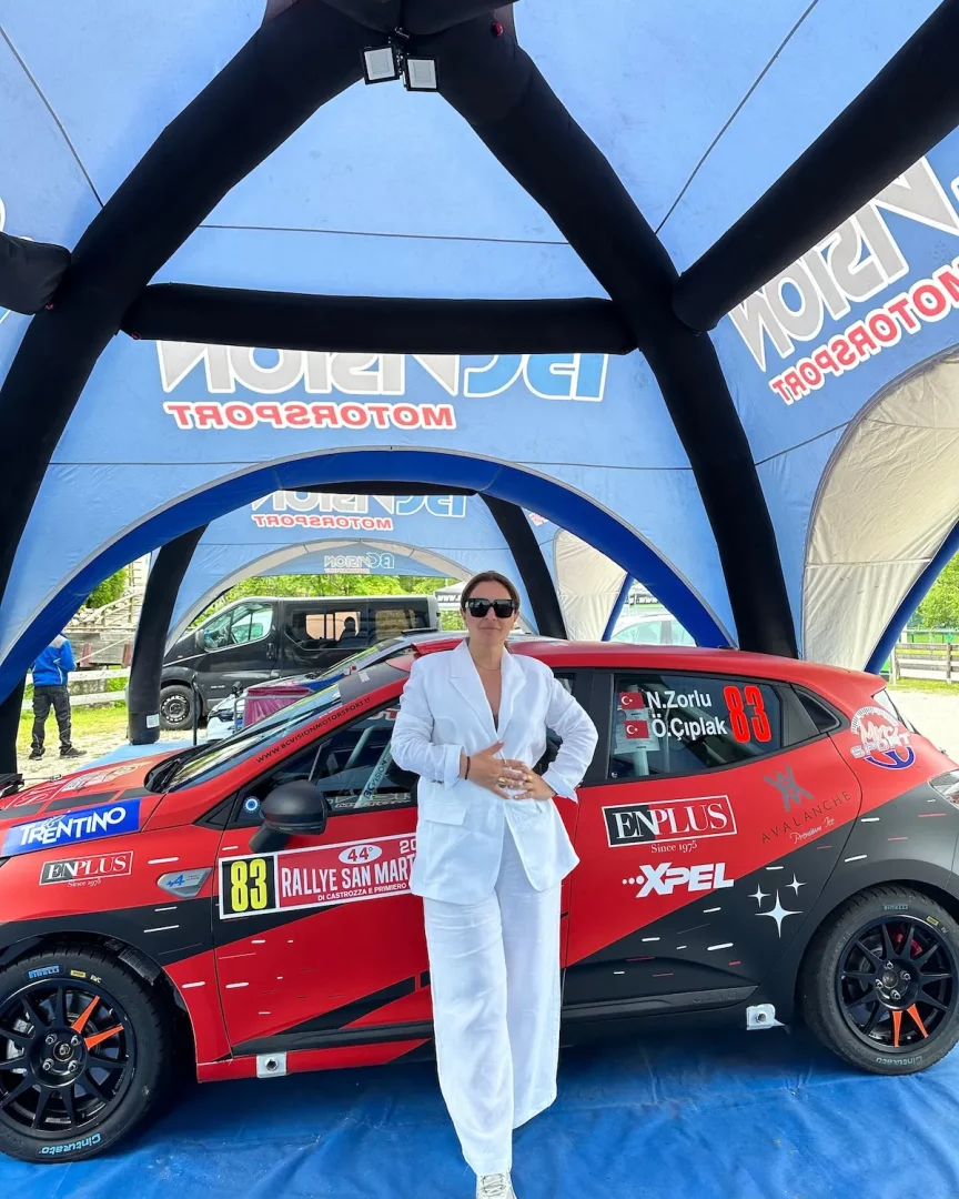 Nazan Zorlu, üzerinde 83 numarası, çeşitli sponsor logoları ve N.Zorlu ile Ö.Çıplak isimlerinin yer aldığı kırmızı renkli bir ralli otomobilinin yanında, beyaz takım elbisesi ve güneş gözlükleriyle dururken, arka planda mavi VISION MOTORSPORT çadırının bir kısmı görülüyor.
