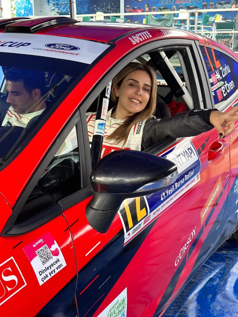 Nazan Zorlu, üzerinde beyaz yarış tulumuyla, 47. Yeşil Bursa Rallisi etiketleri ve sponsor logolarıyla süslenmiş kırmızı-mavi renkli bir ralli arabasının sürücü koltuğundan kameraya gülümseyerek bakıyor; arka planda yolcu koltuğunda bir adam da hafifçe görülüyor.