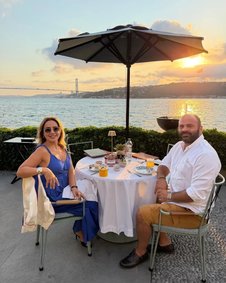 Nazan Zorlu ve yanındaki erkek, İstanbul Boğazı manzaralı bir açık hava restoranında, gün batımının sıcak ışıkları altında şemsiyeli bir masada oturmuş, içecek ve atıştırmalıklarla keyifli vakit geçirirken kameraya gülümser.