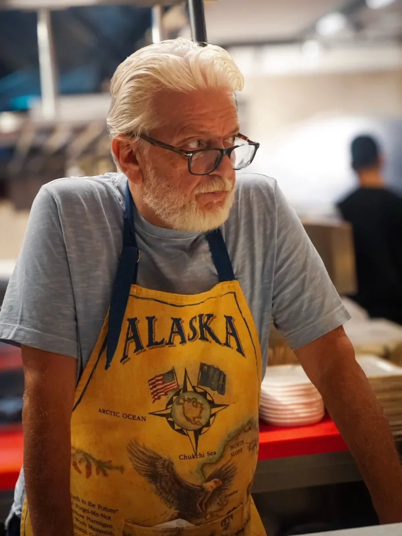 Beyaz saçlı ve sakallı, gözlüklü Uluç Şef, restoran mutfağı ortamında, üzerinde ALASKA yazısı, pusula, Amerikan bayrağı ve kanatlarını açmış bir kartal figürü bulunan sarı önlüğüyle düşünceli bir ifadeyle sağ tarafa bakıyor.