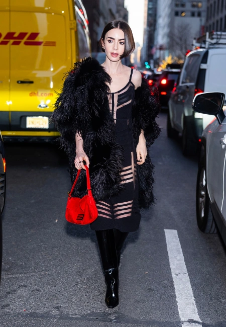 Lily Collins, kısa koyu bob saçları, parlak siyah çizmeleri, tüylü siyah paltosu ve transparan detaylı elbisesiyle kalabalık bir şehir caddesinde yürürken, ikonik Fendi Baguette çantasının canlı kırmızı bir versiyonunu elinde tutuyor.
