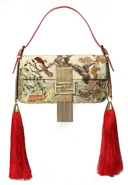 ikonik Fendi Baguette'in 1997 modeli, kuş ve ağaç işlemeleriyle bezeli krem rengi dokuma kumaşı, parlak kırmızı deri sapı, ortasındaki altın renkli FF tokası ve sarkan metalik şerit detayı, her iki yanında sallanan uzun kırmızı püskülleri ve üzerindeki Çince karakterleriyle beyaz bir arka plan üzerinde büyüleyici bir şekilde duruyor.