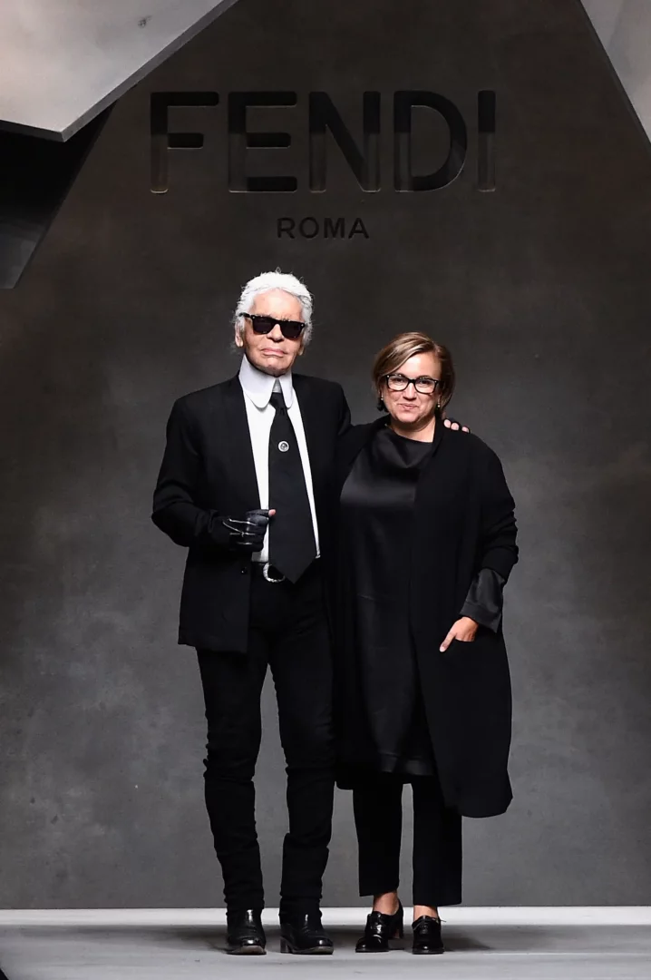 Fendi markasının ikonik isimleri Karl Lagerfeld ve Silvia Venturini Fendi, markanın logosu önünde siyah kıyafetleriyle birlikte poz veriyor; Karl kendine özgü takım elbisesi ve güneş gözlükleriyle, Silvia ise şık bir ceketle zarif bir duruş sergiliyor.