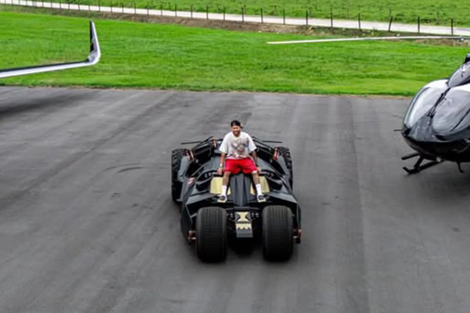 Neymar, asfalt bir zeminde duran, koyu renkli ve fütüristik tasarımlı Batmobile'ın üzerinde beyaz tişörtü ve kırmızı şortuyla otururken görülüyor; arka planda yeşil bir alan, solunda bir uçağın kanadı ve sağında siyah bir helikopter beliriyor.