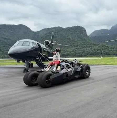 Neymar, özel jetinin önünde, havalimanı pistinde duran, Batman filmlerindeki Tumbler tarzı ikonik Batmobile aracının üzerinde rahat bir şekilde oturmuş, arkasındaki yeşil dağlar ve bulutlu gökyüzü altında poz veriyor.