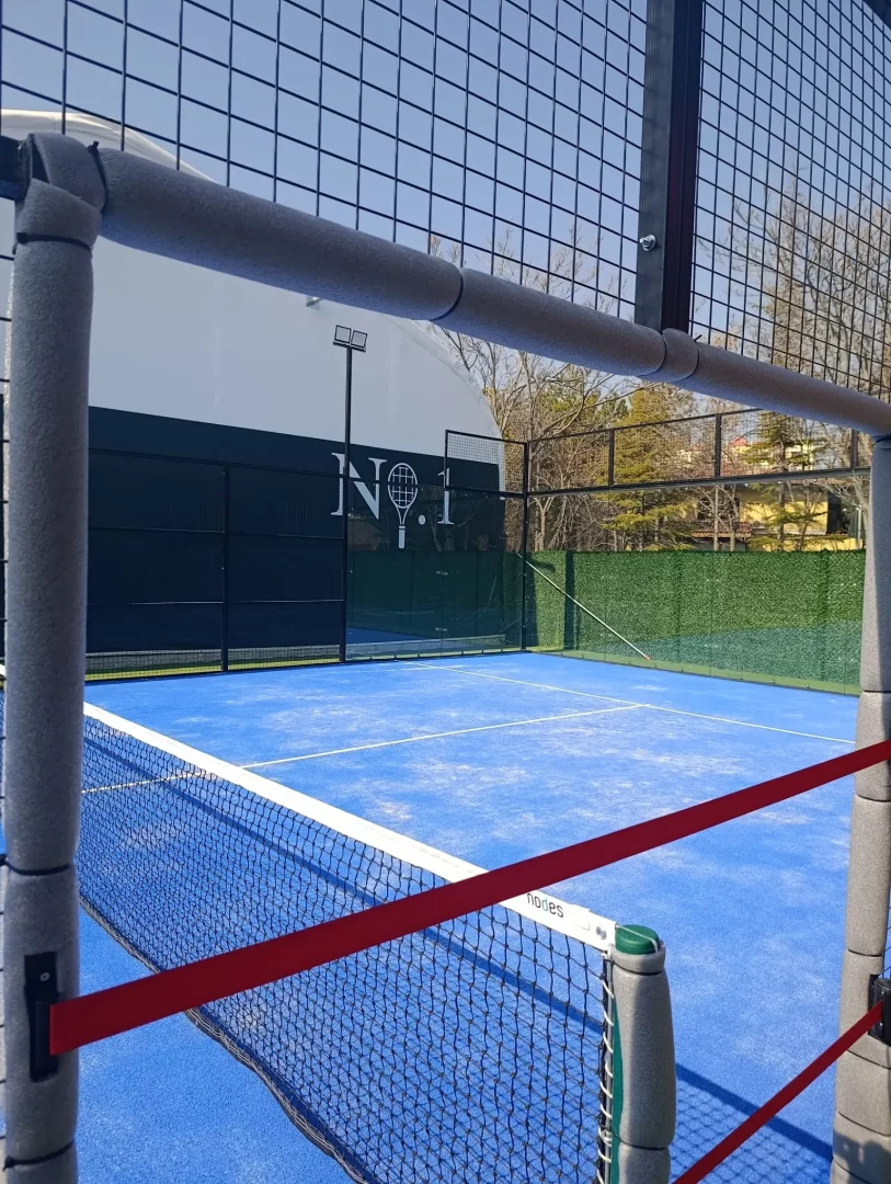 Güneşli bir açık havada, canlı mavi zemini ve beyaz çizgileriyle padel oynamaya hazır, siyah fileli ve direkleri gri koruyucu minderlerle kaplı bir padel kortunun detaylı görünümü; arka planda Nº.1 logosu ve raket sembolü taşıyan beyaz bir tesis binasıyla çevrili bu Ankara padel kortu, aktif bir spor alanını sergiliyor.