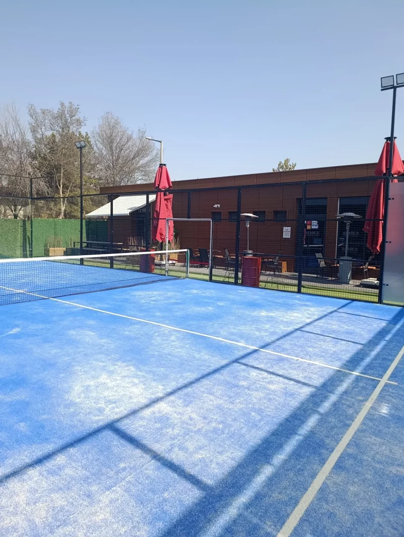 Güneşli berrak bir günde, uzun gölgelerin düştüğü canlı mavi zeminli ve beyaz çizgili padel kortu, arka planda modern bir bina, katlanmış kırmızı şemsiyeler ve ısıtıcılarla donatılmış bir dinlenme alanıyla No1 Padel Ankara'nın açık hava kortlarını sergiliyor.