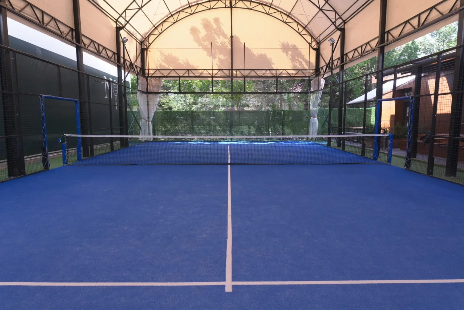 Ankara'daki, üzeri kapalı bir padel kortunun geniş açılı fotoğrafında, canlı mavi zemini beyaz çizgiler ve ortadaki net ile öne çıkarken, metal çerçeveli tavan yapısı ile etrafını saran tel örgülü duvarlar dışarıdaki yeşil ağaçların yansımalarını gösteriyor.