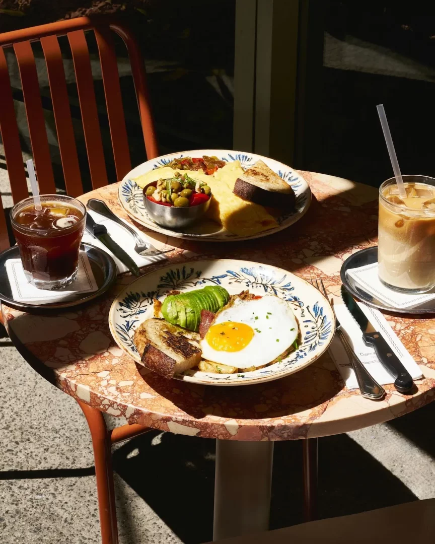 Güneşli bir kafede, benekli mermer bir masanın üzerinde, zeytinli omlet ve avokadolu sahanda yumurta gibi çeşitli kahvaltı veya brunch lezzetlerinin sunulduğu iki tabak, yanında ferahlatıcı buzlu kahve ve buzlu latte ile keyifli bir anı yansıtıyor.