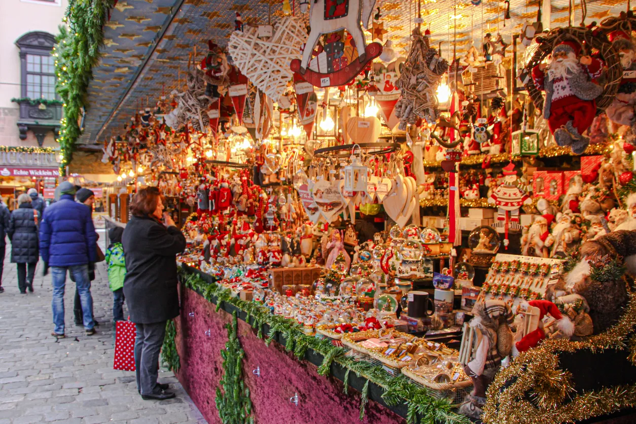 Noel pazarı tezgahında, ahşap at figürleri, kalp şeklinde süslemeler, Noel Baba heykelleri ve ışıl ışıl kar küreleri gibi yüzlerce farklı Noel süslemesinin sergilendiği, kalabalık ve şenlikli bir atmosferde alışveriş yapan insanları gösteren geniş bir görünüm.