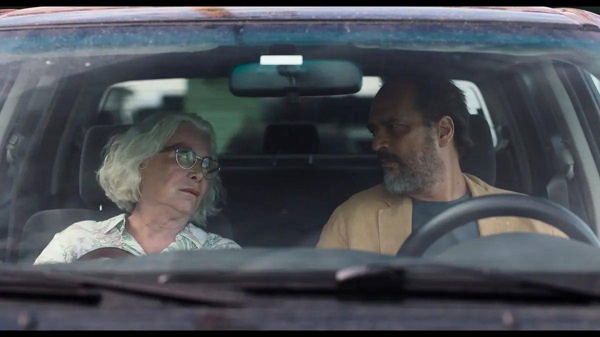 O da Bir Şey mi? film sahnesinde, hafif kirli ön camdan görünen otomobilin içinde, gri saçlı, gözlüklü yaşlı bir kadın ile sakallı bir adam, sanki derin bir sohbete dalmış gibi birbirlerine bakıyorlar.