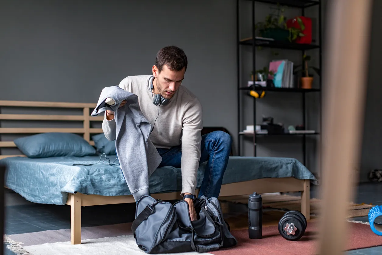 Yatak kenarında oturan, açık renk sweatshirt ve kot pantolon giyen, kulaklığı boynunda olan bir adam, ofis sonrası spora gitmek için gri eşofmanını dikkatle gri spor çantasına yerleştiriyor; yerde bir su şişesi ve dambıl, spor hazırlığını vurguluyor.