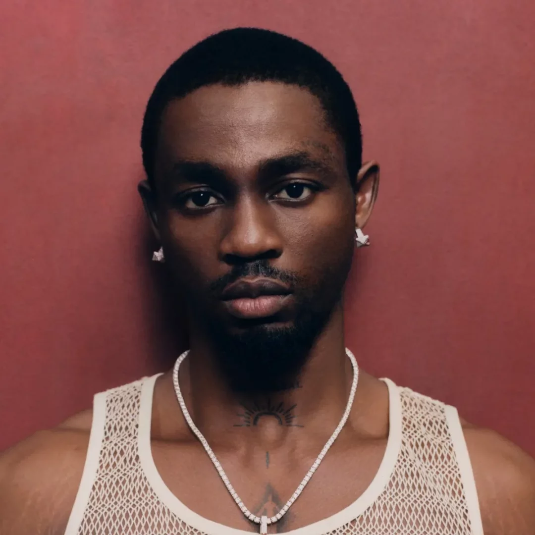 Nijeryalı Afrobeats sanatçısı Omah Lay, İstanbul konseri öncesinde Boy Alone albümünün başarısını temsil eden beyaz file atlet, parlak kolye ve küpelerle, güneş dövmesinin üzerinde ciddi bir ifadeyle kameraya bakıyor.