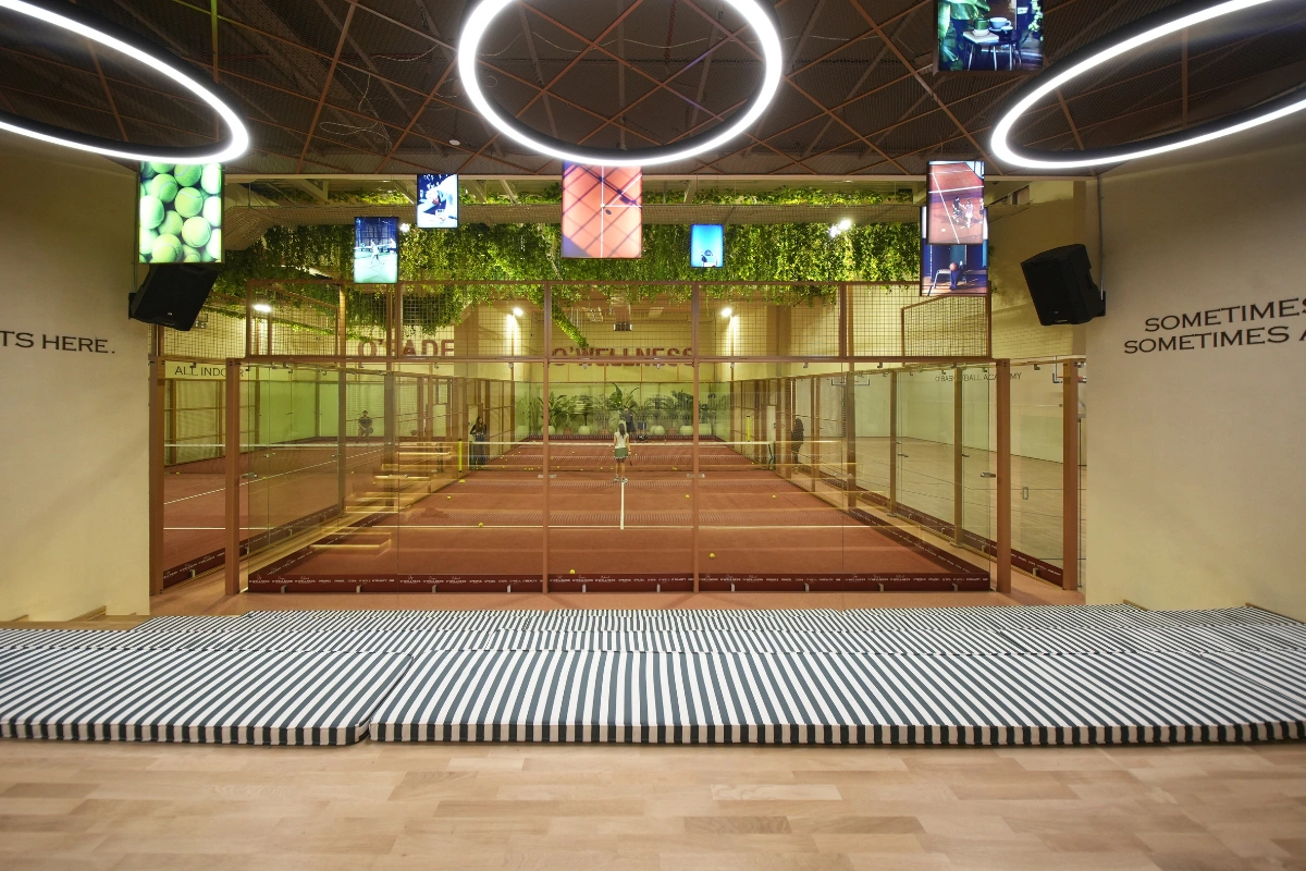 İstanbul Sarıyer'deki modern bir kapalı padel kortu tesisini gösteren bu geniş açılı görselde, cam duvarlarla çevrili kahverengi kortlarda birkaç oyuncu ve etrafa saçılmış sarı toplar görülürken, tavandan sarkan bitki örtüsü, halka şeklindeki ışıklandırmalar ve ön plandaki çizgili minderler mekanı çağdaş ve dinamik bir spor alanı olarak sunmaktadır.