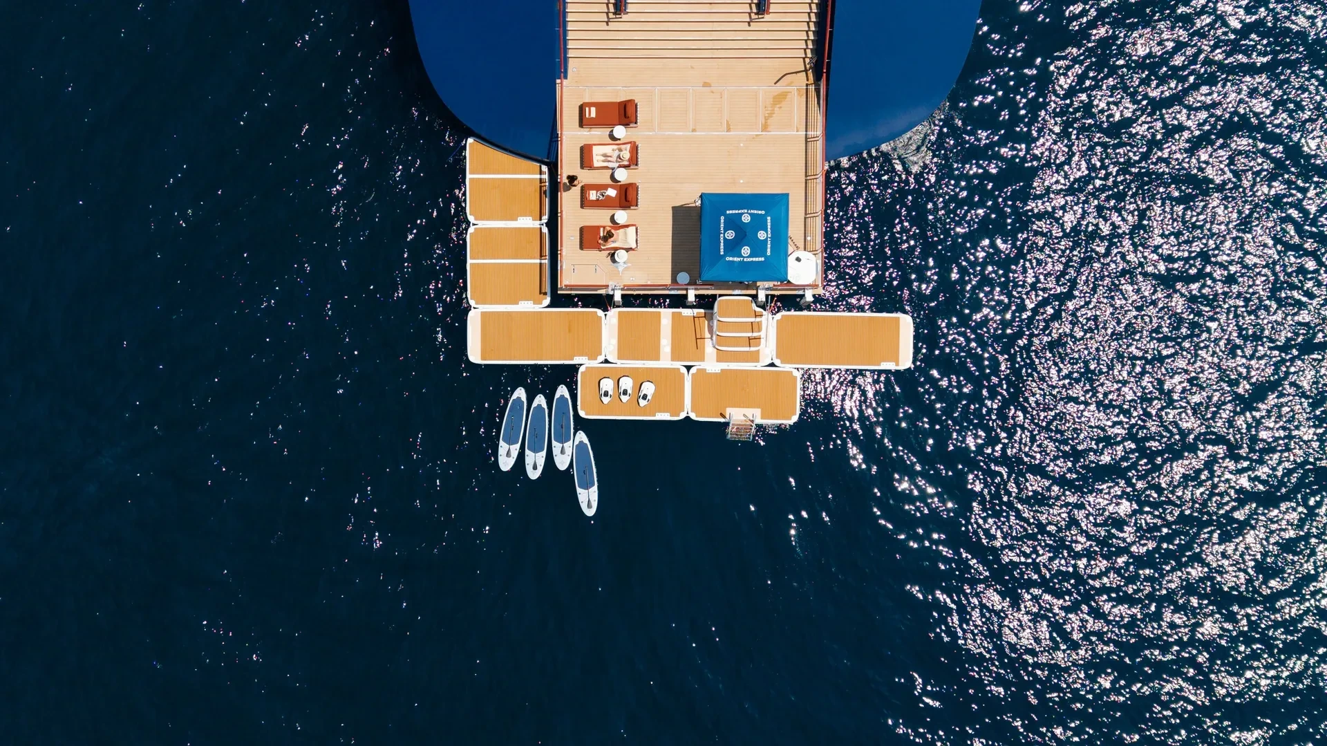 Kuşbakışı görünen lüks bir Orient Express yatının arka güvertesinde, ahşap zeminli geniş platformlarda güneşlenen konuklar ve suya erişim sağlayan sıralanmış paddleboard'lar, modern deniz seyahatinde öne çıkan özel yat deneyimi ve mahremiyet arayışını yansıtıyor.
