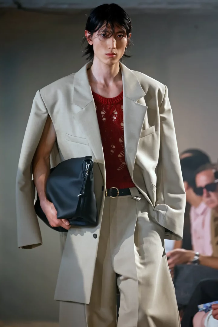 Podyumda yürüyen, bej renkte, omuzları düşürülmüş ve kolları geniş kesimli bir oversize blazer ceket giyen model, içine giydiği kırmızı delikli üstü ve elindeki siyah çantasıyla modern ve kendine özgü bir tarzı yansıtıyor.