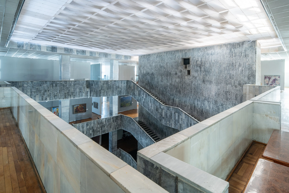 Çok katlı, geniş bir modernist iç mekanın gösterildiği görselde, Taşkent'in 1966 depremi sonrası yeniden yapılanma sürecinde inşa edilen ve UNESCO Dünya Mirası Listesi'ne giren mimari eserlerden birinin gri taş ve mermer kaplı duvarları, cesur geometrik çizgilerle tasarlanmış merdivenleri ve sanatsal tabloların sergilendiği alanları belirgin bir şekilde öne çıkıyor.