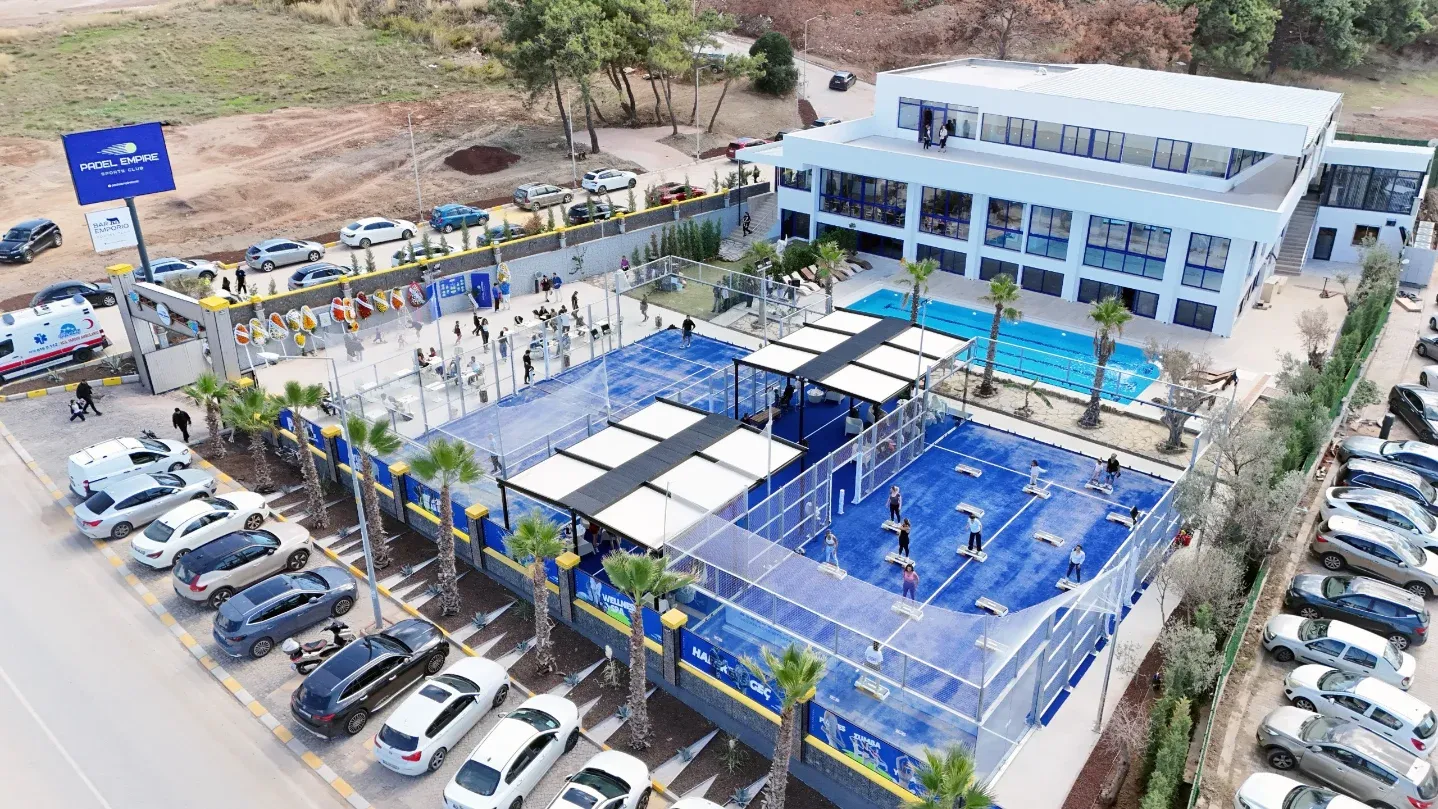 İzmir'deki Padel Empire Sports Club'ın kuşbakışı görüntüsü, çok sayıda mavi padel kortu, modern bir kulüp binası, yüzme havuzu ve kalabalık otoparkıyla canlı bir spor kompleksini sergiliyor.