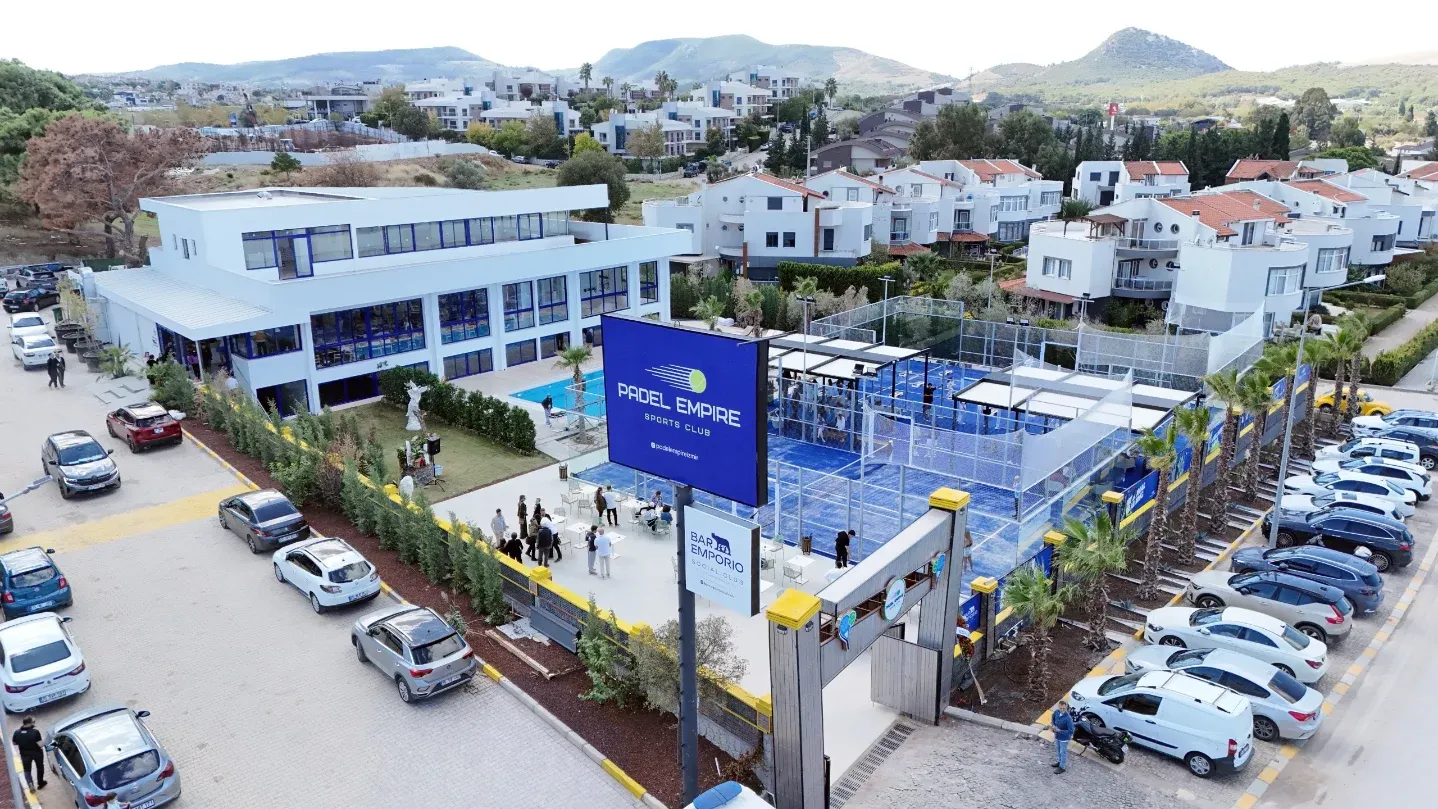 Padel Empire Sports Club'ın havadan çekilmiş bu geniş görüntüsünde, modern tasarımlı ana bina, canlı mavi renkli padel kortları, kalabalık bir sosyal alan ve çevredeki konutlarla birlikte, İzmir'deki tesisin aktif ve davetkar atmosferi öne çıkıyor.