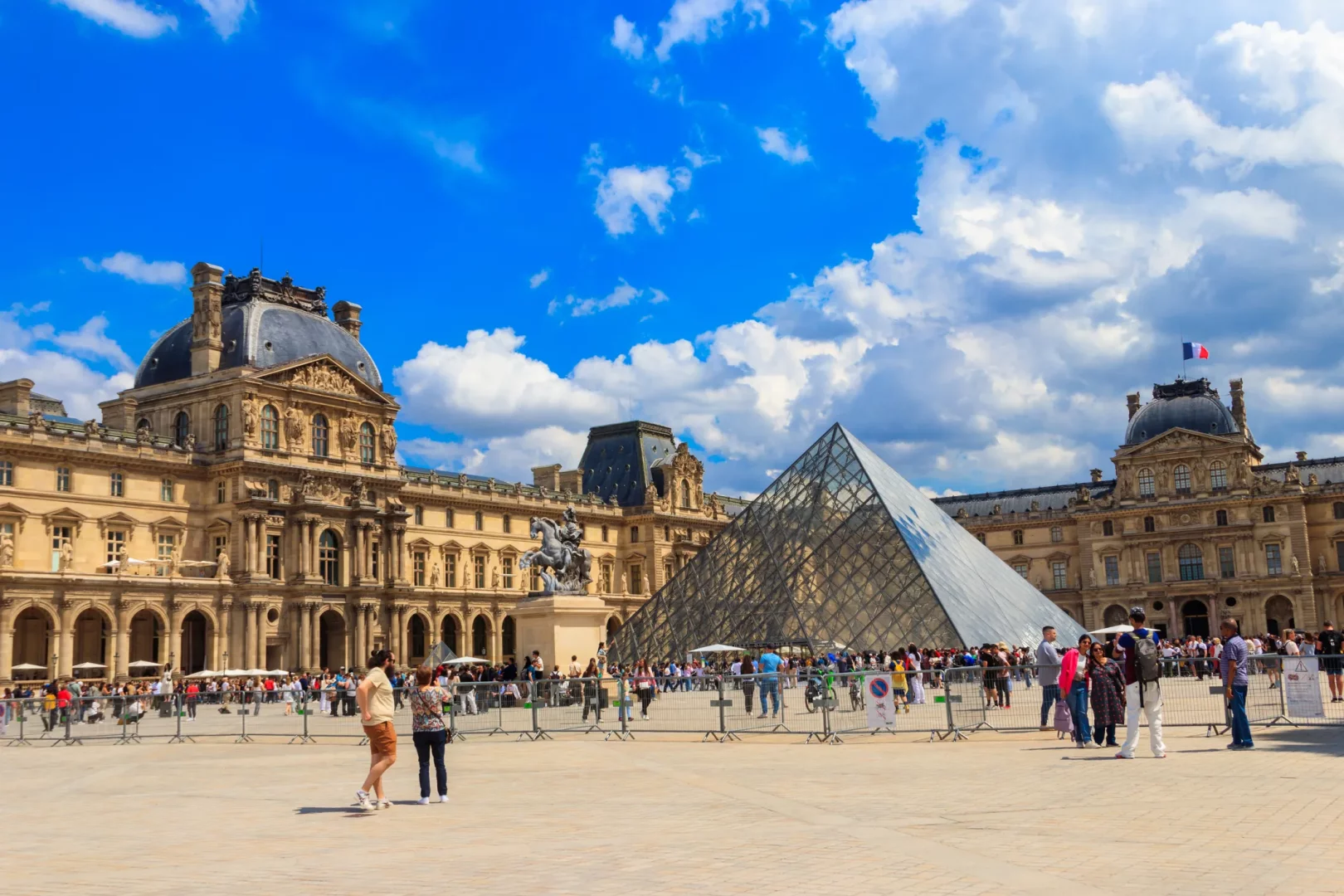 Paris'in dünyaca ünlü sanat ve tarih merkezi Louvre Müzesi'nin geniş avlusunda, görkemli Barok mimarili tarihi saray binalarının arasında göğe uzanan ikonik cam piramit, çatıdaki Fransız bayrağı ve güneşli bir günde müzeyi ziyaret eden kalabalık turistlerin oluşturduğu hareketli bir görüntü.