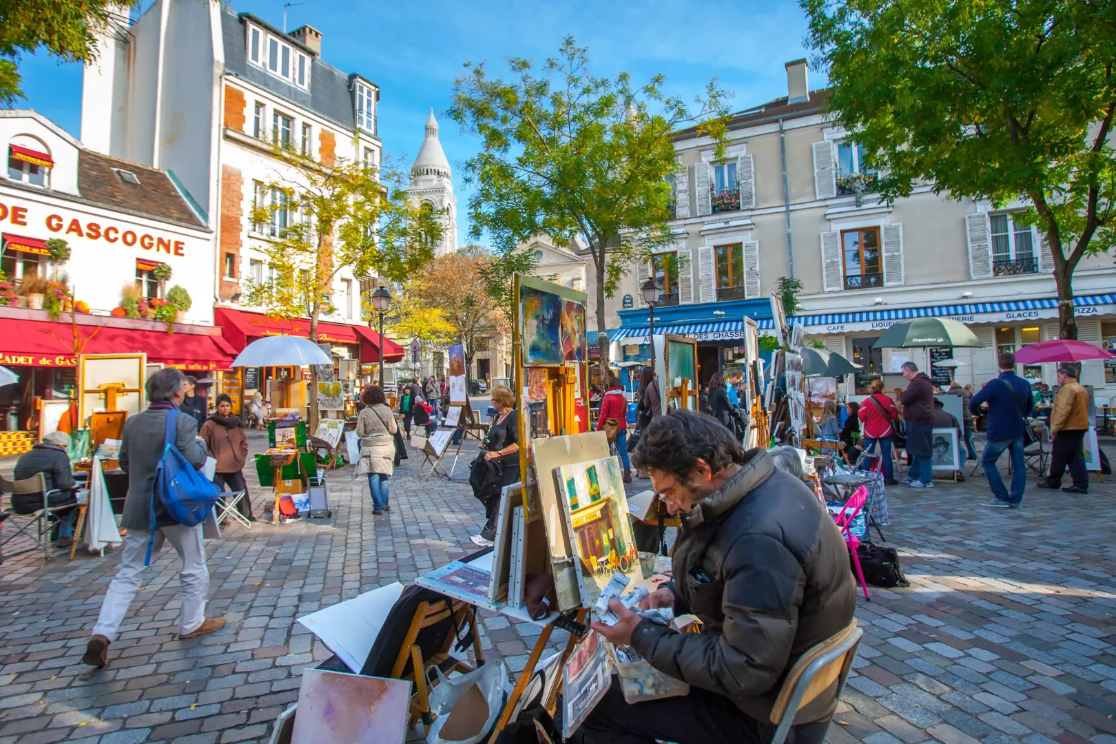 Paris'in hareketli Montmartre bölgesindeki tarihi Place du Tertre Meydanı'nda, Sacré-Cœur Bazilikası'nın bembeyaz kubbesinin uzaktan göründüğü pırıl pırıl bir sonbahar gününde, birçok sanatçı şövalelerinin başında eserlerini sergilerken, ön planda dikkatle tablosuna odaklanmış bir ressam ve etrafta dolaşan ziyaretçiler canlı bir atmosfer yaratıyor.
