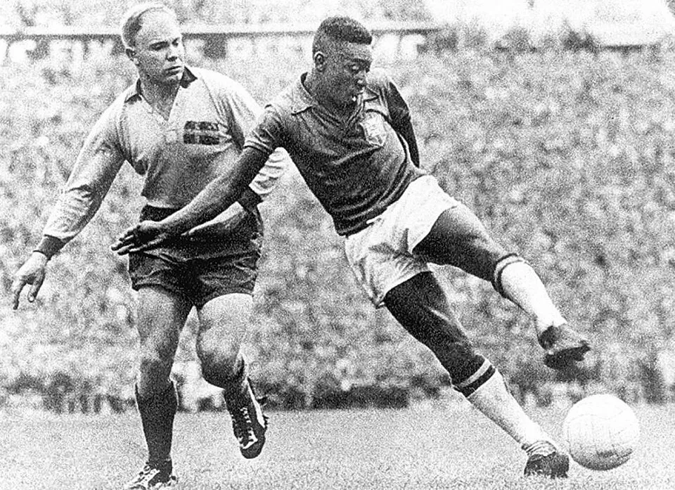 Siyah beyaz bu karede, futbol efsanesi Pelé, 1958 Dünya Kupası İsveç'teki bir maçta koyu renkli Brezilya forması ve açık renkli şortuyla topa sağ ayağıyla ustaca vuruyor; hemen arkasında açık renkli formasıyla bir hakem oyunu dikkatle takip ederken, bulanık tribünler izleyici kalabalığıyla dolu.