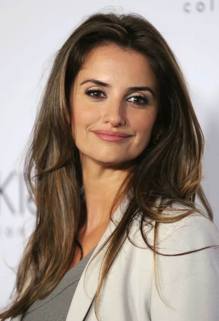 Penelope Cruz, esmerlere yakışan uzun, dalgalı, kahverengi saçları ve hafif karamel balyajlarıyla gülümseyerek kameraya bakıyor; bu görünüm, esmerler için ideal saç renkleri seçeneklerinden birini başarıyla sergiliyor.