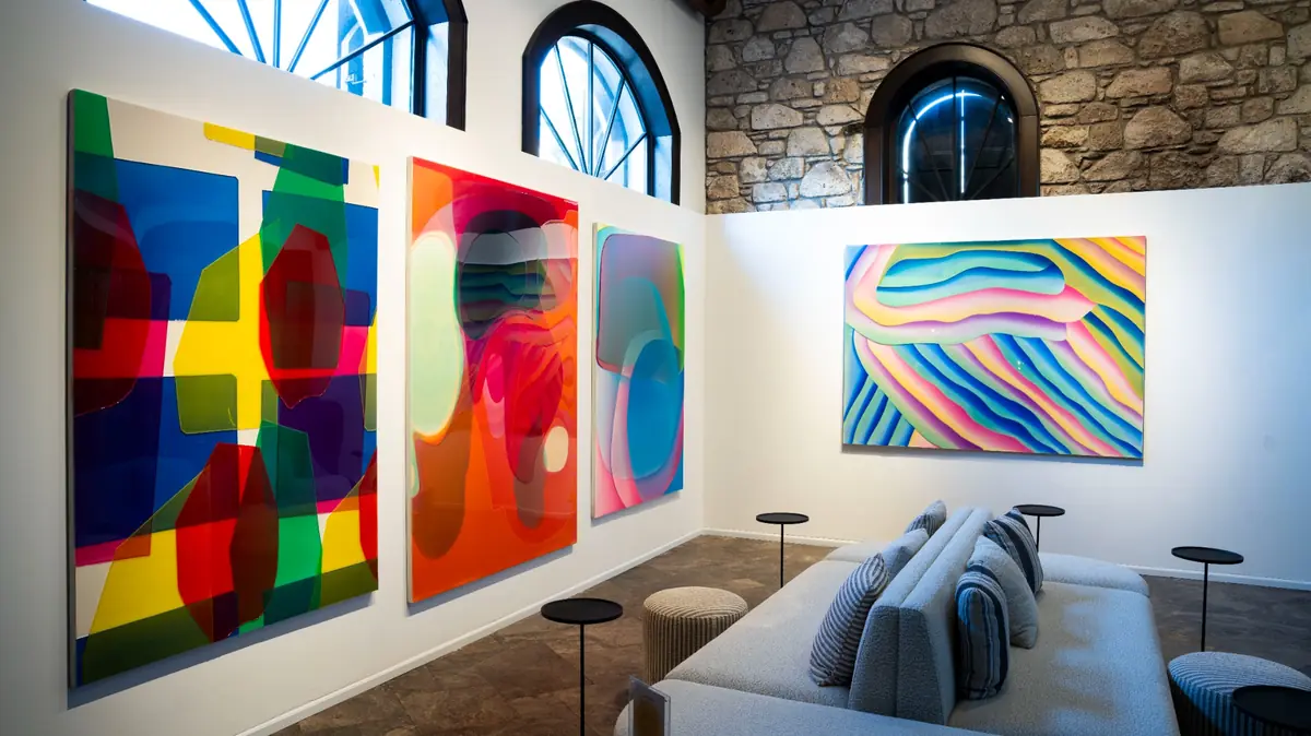 Alaçatı'daki LS Contemporary'de sergilenen Peter Zimmermann'ın imzası niteliğindeki canlı renkli ve katmanlı soyut eserlerinin beyaz duvarlarda sergilendiği, kemerli pencereleri ve taş duvar detaylarıyla modern bir sanat galerisi iç mekanında, ön planda konforlu bir oturma grubunun ve yan sehpaların yer aldığı geniş bir genel görünüm.